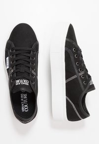 Černé nízké plátěné tenisky s tlustými bílými podrážkami, bílým prošíváním, černými tkaničkami a logem "Versace Jeans Couture" na jazyku a stranách.