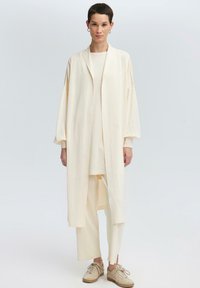 Cardigan lungo di colore crema, aperto davanti, con spalle scese, indossato sopra un top abbinato e pantaloni cropped. Sneakers beige completano l'outfit.