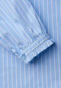 Chemise en coton bleu clair à rayures blanches, avec un poignet froncé, une fermeture à boutons et une texture subtile.