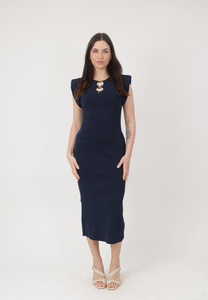 Frau mit langen dunklen Haaren, die ein ärmelloses Midikleid in Marineblau und beige Sandalen mit Absatz trägt, steht vor einem schlichten hellen Hintergrund.