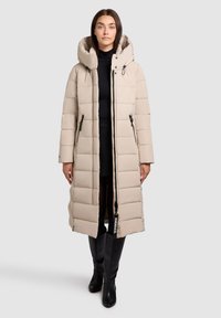 Dlhý béžový vatovaný kabát s vysokým golierom, vreckami na zips a čiernym jazdcom na zips. Štruktúrovaný prešívaný vzor. Nosený na čiernom outfite.