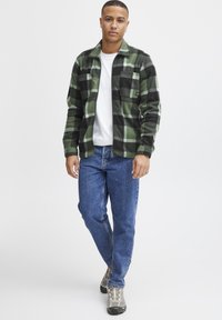 Groen-en-zwarte geruite flanellen shirt met een wit t-shirt eronder, gecombineerd met blauwe spijkerbroek en grijze wandelschoenen.