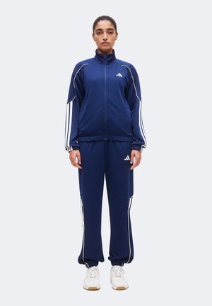 Kobieta stojąca twarzą do przodu, ubrana w granatowy dres Adidas z białymi paskami i białe trampki na jednolitym jasnym tle.