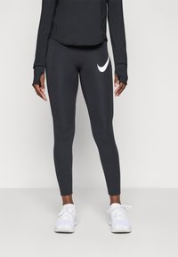 Person som bär svarta Nike leggings med vitt swoosh-logotyp, svart långärmad tröja och vita sportskor, stående mot en enkel bakgrund.
