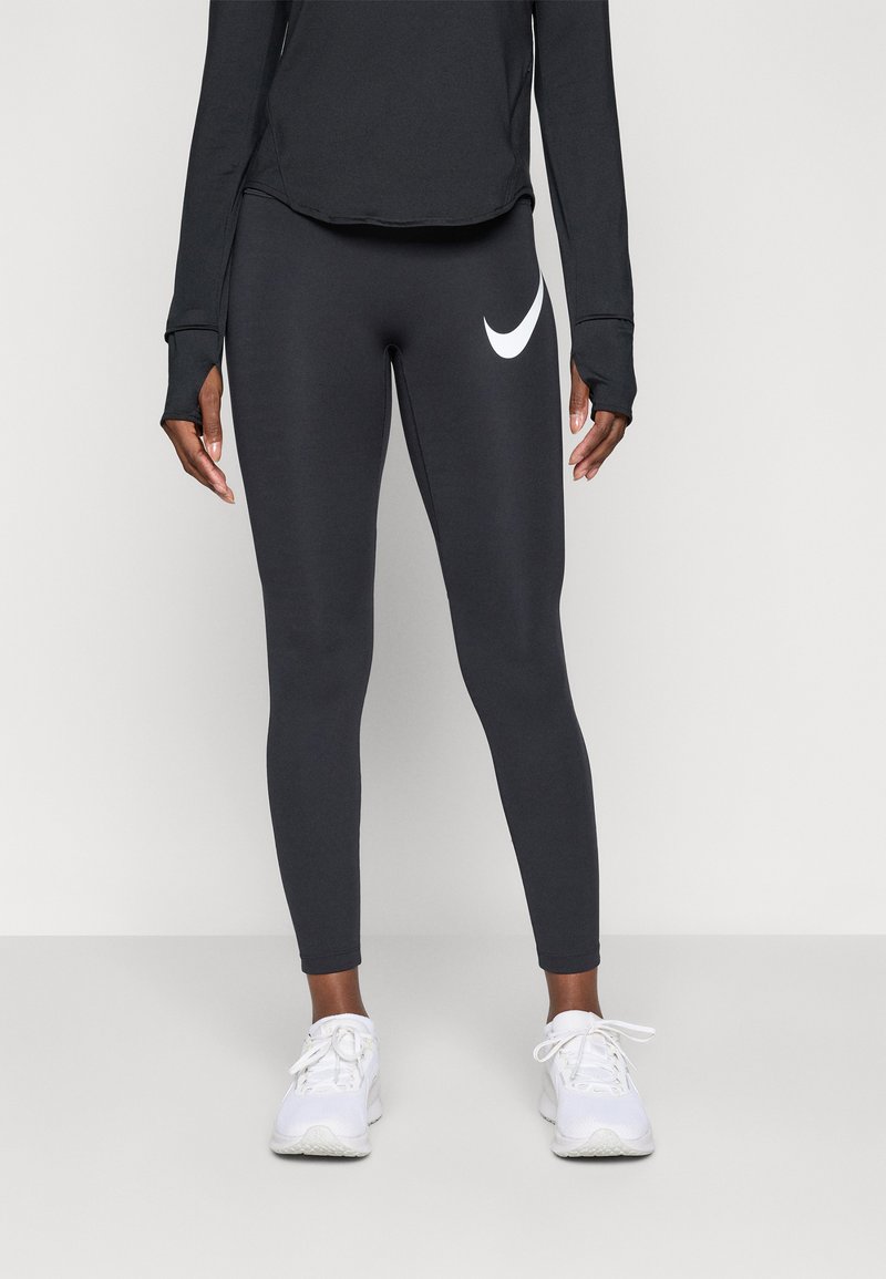 Person som bär svarta Nike leggings med vitt swoosh-logotyp, svart långärmad tröja och vita sportskor, stående mot en enkel bakgrund.