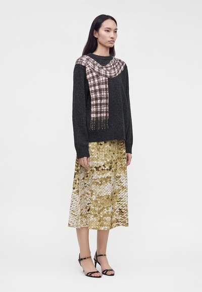 Femme debout portant un pull noir avec une écharpe à carreaux roses et une jupe midi dorée à motifs, avec des talons ouverts noirs.