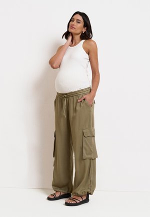 Schwangere Frau trägt ein weißes Tanktop, lockere olivgrüne Cargohosen und schwarze Sandalen, steht mit einer Hand am Hals und einer in der Tasche.