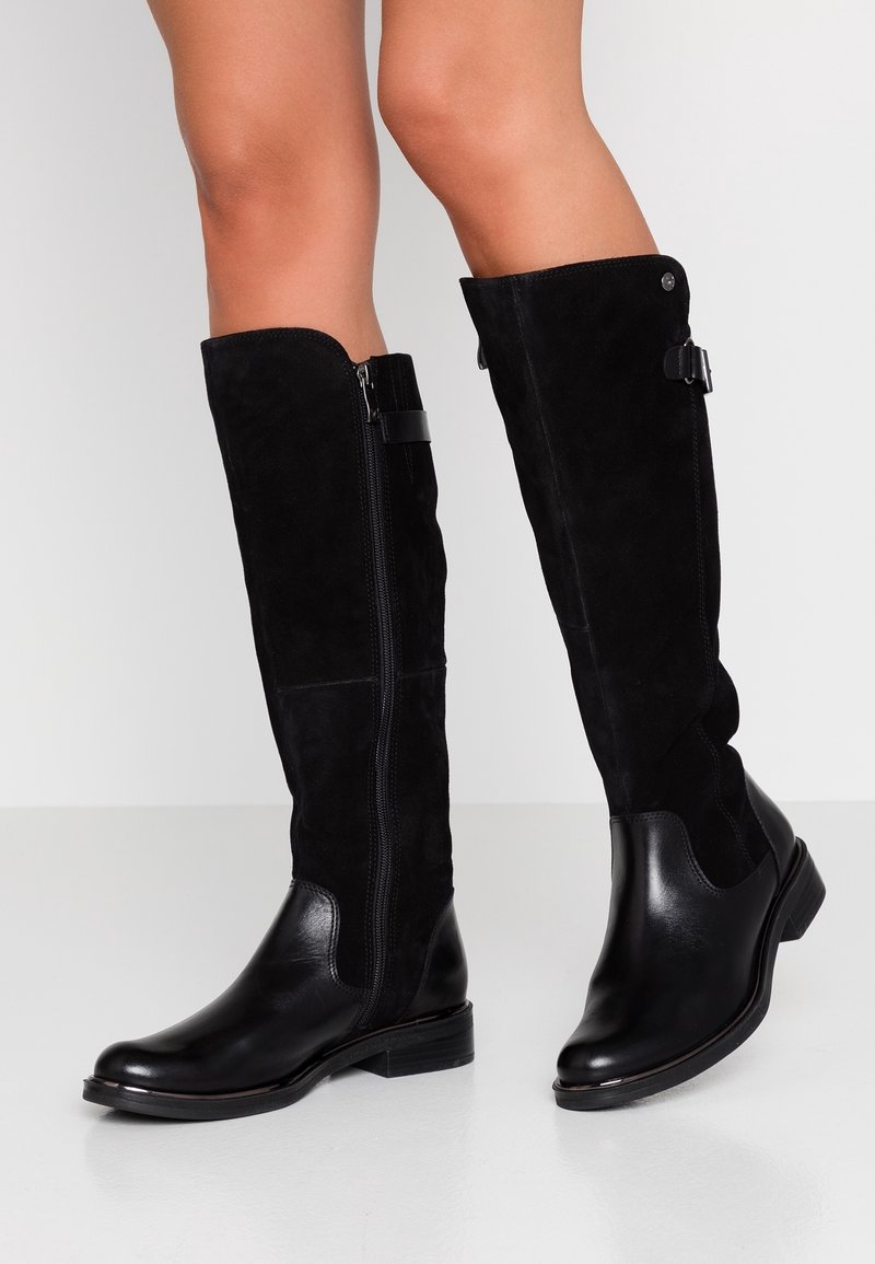 Bottes noires montant jusqu'aux genoux en cuir lisse et en suède texturé, avec une fermeture éclair latérale et des accents de sangle décoratifs. Talons plats.