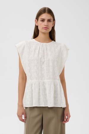 Mujer joven que lleva una blusa blanca sin mangas con estampados florales y pantalones anchos beige, de pie frente a un fondo liso.