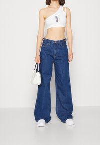Top corto blanco de un solo hombro, jeans anchos azul con detalles de botones, combinados con un pequeño bolso blanco y zapatillas blancas.