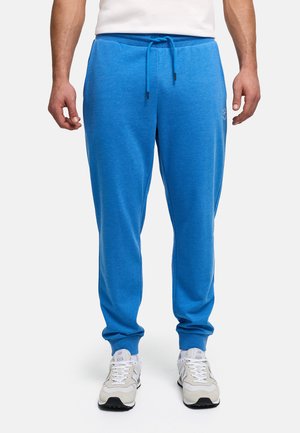 Bărbat purtând pantaloni de trening albastru aprins cu șnur și pantofi sport albi, stând pe un fundal alb simplu.