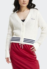 Mulher vestindo um cardigan branco de malha com botões e riscas azuis, com uma saia vermelha da Adidas com riscas brancas, posando com uma mão na cintura.