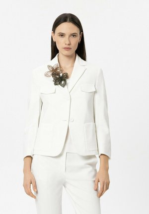 Frau trägt einen weißen Anzug mit aufgesetzten Taschen und dekorativen Drahtblumenbroschen am linken Revers, steht vor einem einfarbigen Hintergrund.