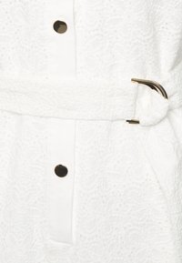 White dress featuring textured lace with a floral pattern, a front button closure, and a matching belt with a gold buckle.