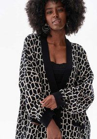 Zwarte en crèmekleurige cardigan met een geometrisch honingraatpatroon, open voorkant, lange mouwen en ribgebreide manchetten, gemaakt van een gestructureerd breimateriaal.