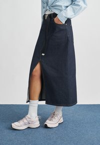 Μακριά φούστα από σκούρο τζιν με εμπρόσθια σχισμή, πλαϊνές τσέπες και μαύρη ζώνη. Συνδυασμένη με αθλητικά παπούτσια σε ανοιχτό ροζ και λευκές κάλτσες.