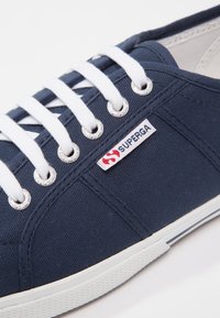 Modré plátěné tenisky s bílými šňůrkami, stříbrnými oky a gumovou podrážkou. Na boku se nachází červenobílá visačka loga Superga.