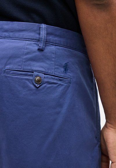 Κοντινό πλάνο μπλε παντελονιού chino με τσέπη στο πίσω μέρος με κουμπί και κεντημένο λογότυπο αλόγου στο δεξί ισχίο, φορεμένο από άτομο με σκουρόχρωμο δέρμα.