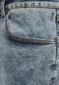 Tissu en denim bleu avec une texture délavée et usée. Comprend une poche latérale avec des coutures orange et des détails en métal.