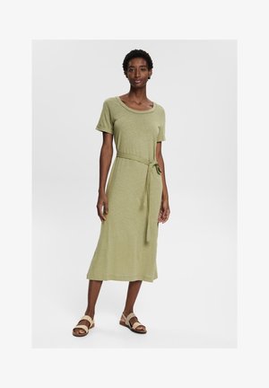 Abito verde oliva, lungo fino al ginocchio, realizzato in tessuto morbido. Presenta maniche corte, scollatura a barchetta e un dettaglio di vita legato. Abbinato a sandali beige.