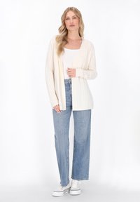 Cremefarvet kabelstrik åben cardigan med rib strikkede manchetter, båret over en hvid tanktop og parret med lyseblå brede bukser og hvide sneakers.