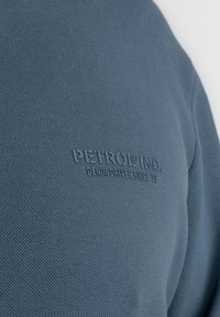 Blauwe gemerceriseerde stof met een subtiele reliëflogo dat "PETROL IND. DENIM DRIVEN SINCE '89" zegt, gelegen aan de linkerkant.