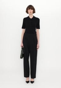 Polo noir à manches courtes avec des boutons, associé à un pantalon large noir taille haute. Porte un sac à main noir texturé et des chaussures noires à bout pointu.