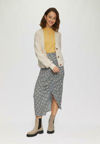 Beige Strickjacke mit großen Knöpfen, über einem senfgelben Oberteil und einem gemusterten Midirock getragen. Graue Knöchelschafter mit schwarzen Details.