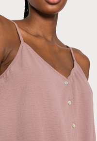 Haut sans manches rose clair avec un col en V, doté d'une fermeture devant à boutons. Confectionné dans un tissu texturé avec de fines bretelles.