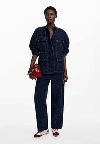 Tumesinist teksast jakk kahe klapiga taskuga, millele on lisatud sobivad laiade säärtega püksid. Modell hoiab punast kotti ning kannab punaseid ja valgeid tossusid.