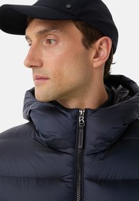 Navy puffer jacket featuring a high collar, zipper front with branded pull, and smooth, glossy texture. Paired with a matching cap.