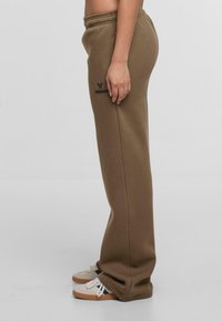 Braune Sweatpants mit weichem Stoff, ausgestattet mit einem verstellbaren Bund und einem gestickten Logo am linken Oberschenkel. Getragen mit weißen Sneakers.