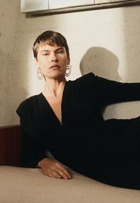Black fabric dress with a plunging neckline, smooth texture, and shaped sleeves. Large circular earrings and a neutral backdrop.