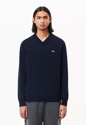Suéter de punto de color azul marino con cuello en V, presentado con un logo verde de Lacoste en el lado izquierdo del pecho y mangas largas con puños acanalados.
