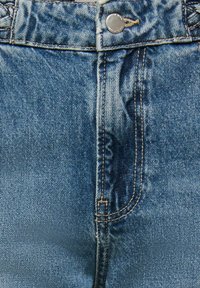 Detailní záběr modrých džínů z denimu se zapínáním na knoflíky, poutky na pásek a švy na předním kapse.