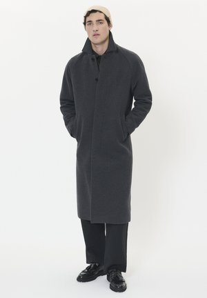 Dunkelgrauer Wollmantel mit einem Kragen, Knopfverschluss und Seitentaschen, kombiniert mit schwarzen Hosen und Schuhen.