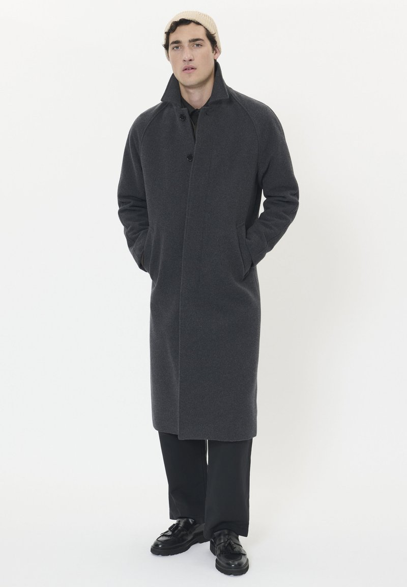Cappotto di lana grigio scuro con collo a revers, chiusura a bottoni e tasche laterali, abbinato a pantaloni neri e scarpe.