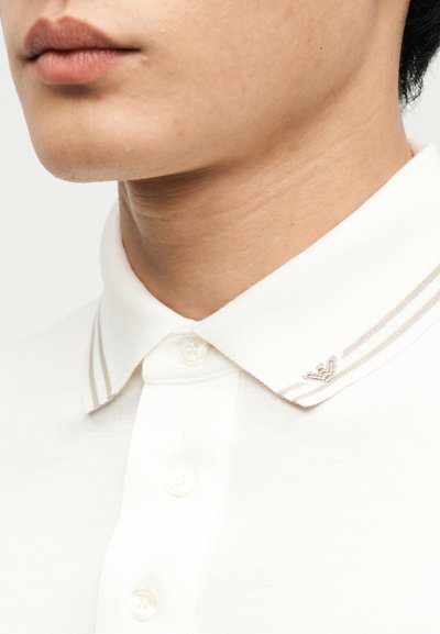 Κοντινό πλάνο του κάτω μέρους του προσώπου και του λαιμού ενός ατόμου που φοράει άσπρο πουκάμισο με λεπτές ρίγες και μικρό κεντημένο λογότυπο γιακά.