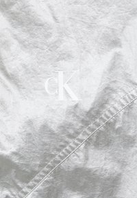 Srebrny materiał o pomarszczonej fakturze z białym logo "cK" na środku. Szczegół szwu przebiegający ukośnie przez powierzchnię.