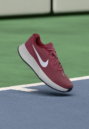 Vínová bežecká obuv Nike s bielym logom, vo vzduchu nad modro-zeleným tenisovým ihriskom pri hranici.