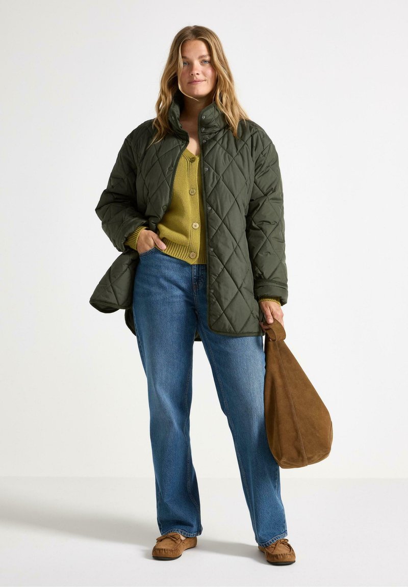 Donna in una giacca imbottita verde sopra un maglione senape, jeans blu e borsa marrone, in piedi su uno sfondo chiaro, sorridendo con sicurezza.