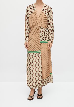 Femme portant une longue robe à motifs avec des dessins géométriques beige et marron, une bordure verte, des manches longues et des sandales à talons noires à brides.