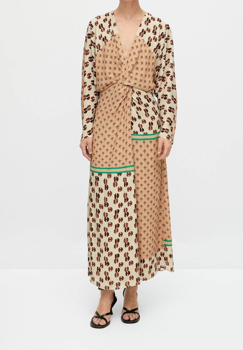 Femme portant une longue robe à motifs avec des dessins géométriques beige et marron, une bordure verte, des manches longues et des sandales à talons noires à brides.