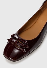 Ballerines en cuir verni bordeaux avec deux petits nœuds au niveau des orteils, bout rond et doublure intérieure lisse.