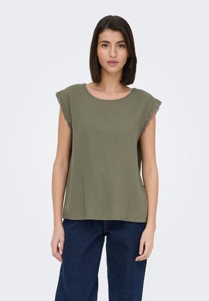 Femme aux cheveux raides et foncés portant un haut sans manches vert olive avec des bordures en dentelle et un jean bleu foncé, debout devant un fond uni.