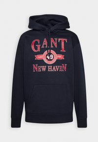 Sudadera con capucha en color navy, con un bolsillo frontal, que presenta un texto en rojo intenso: "GANT 49 NEW HAVEN" y un logo redondo. Material de algodón, corte estándar.