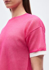 Γυναίκα που φοράει φωτεινό ροζ διαφανές τοπ με κοντά μανίκια πάνω από λευκό πουκάμισο, εμφανιζόμενη από το στήθος ως το λαιμό πάνω σε απλό φόντο.