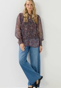 Doorzichtige bloemenblouse met lange mouwen en een strikhalslijn, voorzien van een meerkleurig patroon op een donkere achtergrond, gecombineerd met wijde jeans.