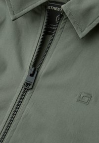 Detailní záběr přední části olivově zelené bundy s černým zipem částečně otevřeným, s límcem a malým vyšitým logem na hrudi.