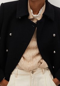 Abrigo negro cruzado con botones plateados llevado sobre un suéter beige de punto trenzado y pantalones crema plisados de talle alto.